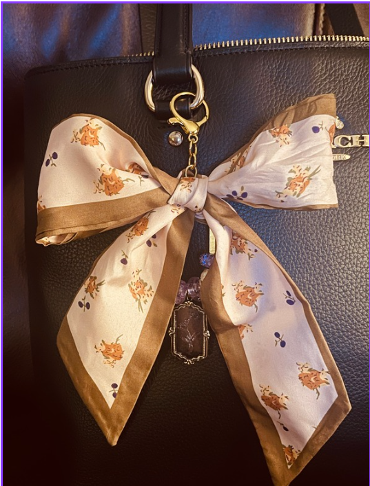 Ivory floral bow bag charm with tan trim and dangling pendant on a black handbag